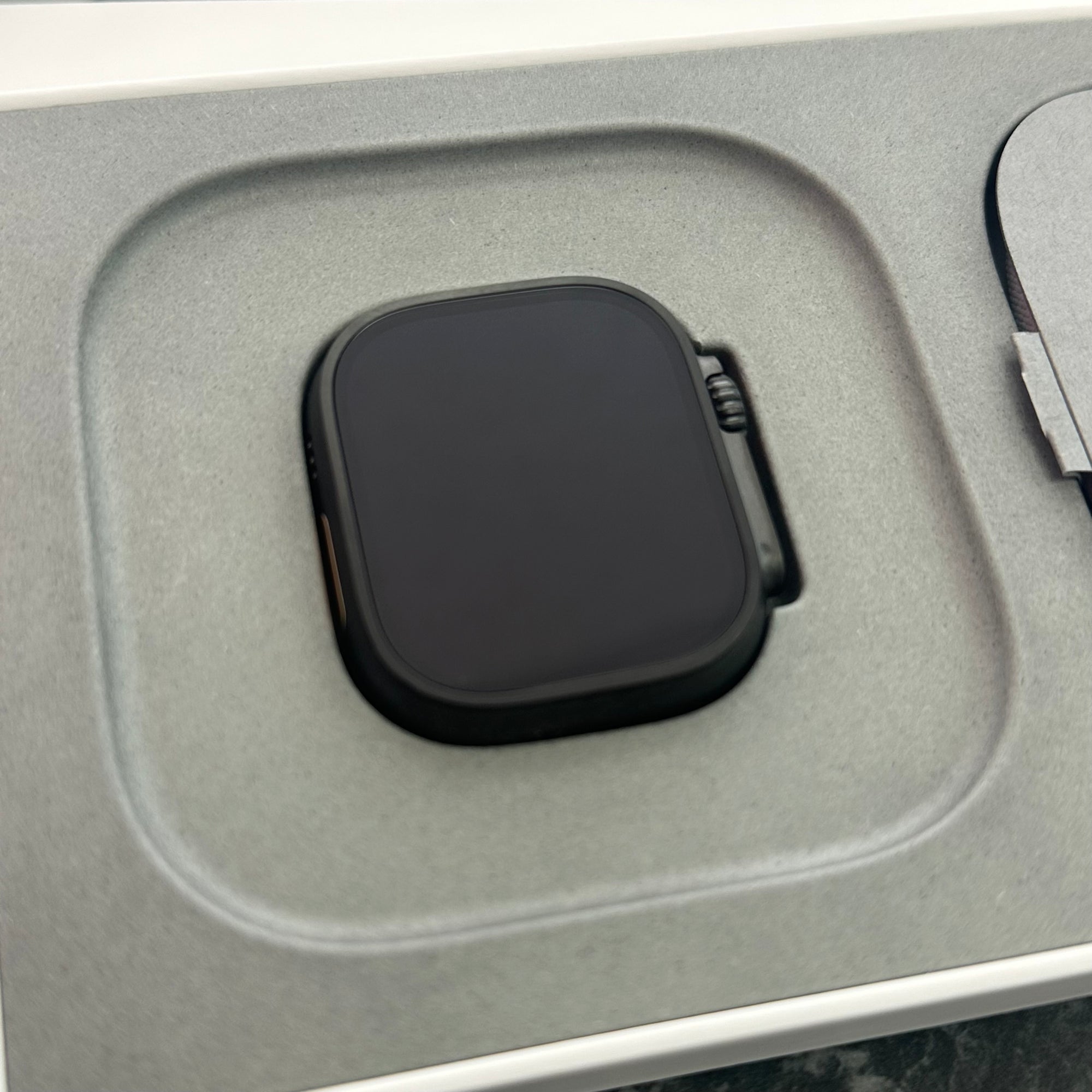 A black apple ultra smartwatch with a square screen and side button, resting in a gray foam insert tray inside a white box.