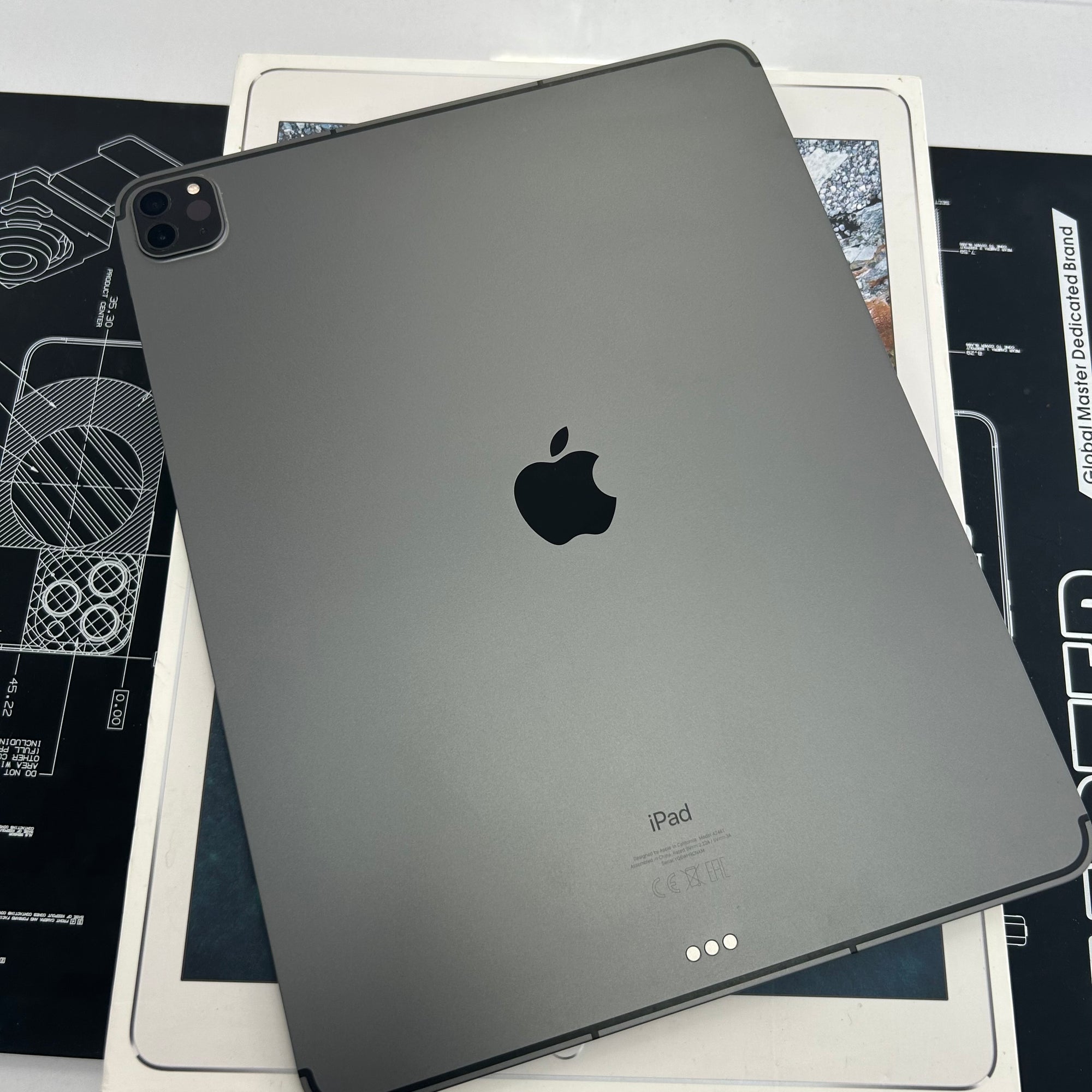 A dark gray Apple iPad shown from the back, resting on top of its white retail box and a black technical schematic mat.