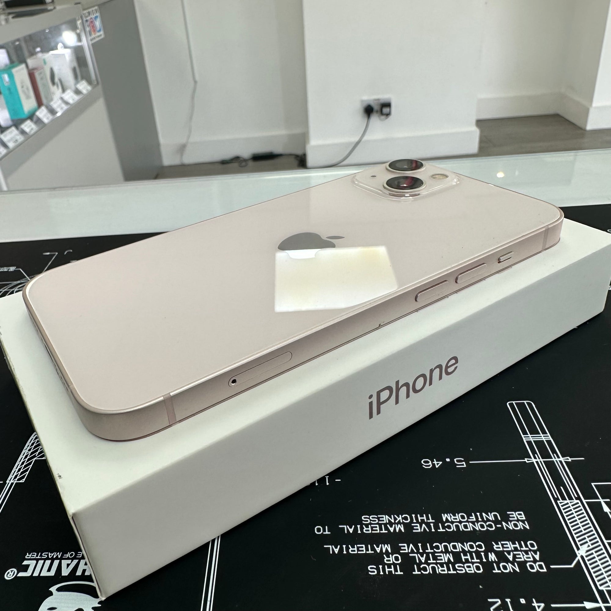 A rose gold iPhone placed on top of its white Apple box, photographed indoors on a black printed surface with a glass table above it.