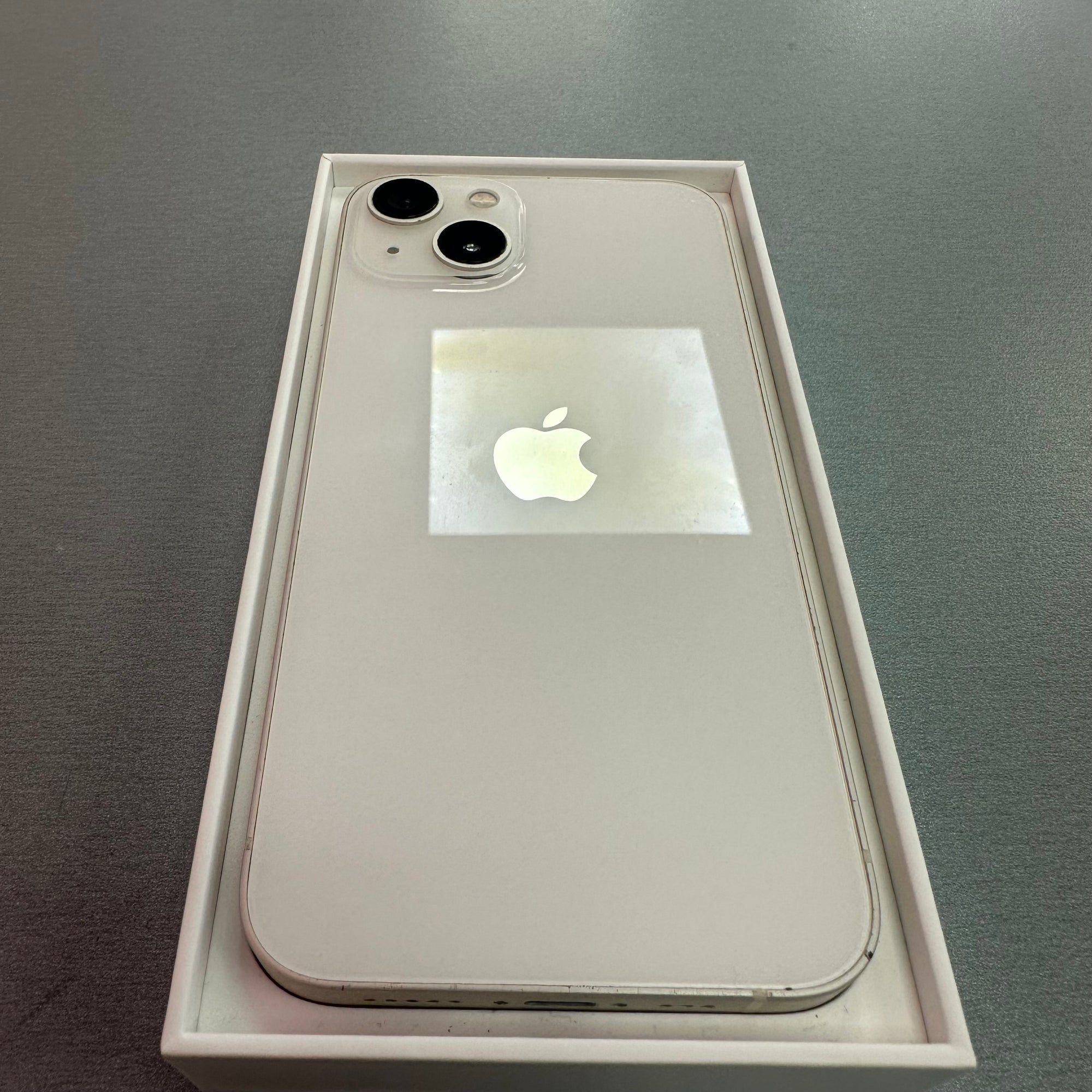 An Apple iPhone in a light gold or pale pinkish-gold finish, shown inside its original retail box on a gray surface. The back of the phone has a protective reflective sticker with the Apple logo, and two rear cameras are visible.
