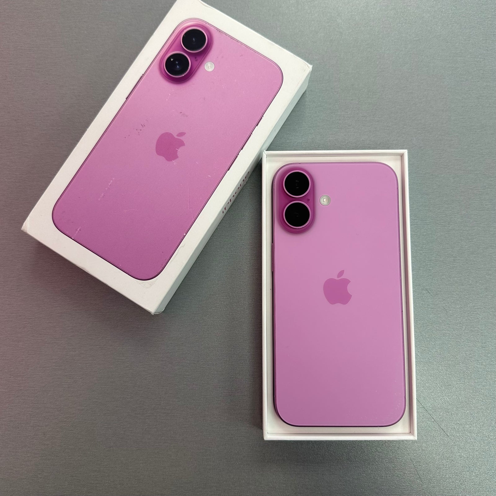 A pink Apple iPhone 16 shown from the back, placed inside its white retail box on a gray surface. The phone has a matte pink finish with a matching pink camera ring and the Apple logo is visible.