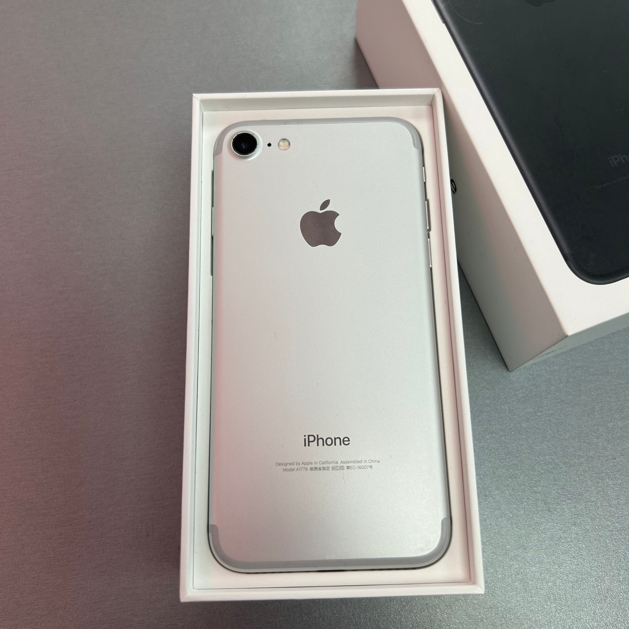 An Apple iPhone 7 in silver color, displayed in its open box next to the box's packaging.