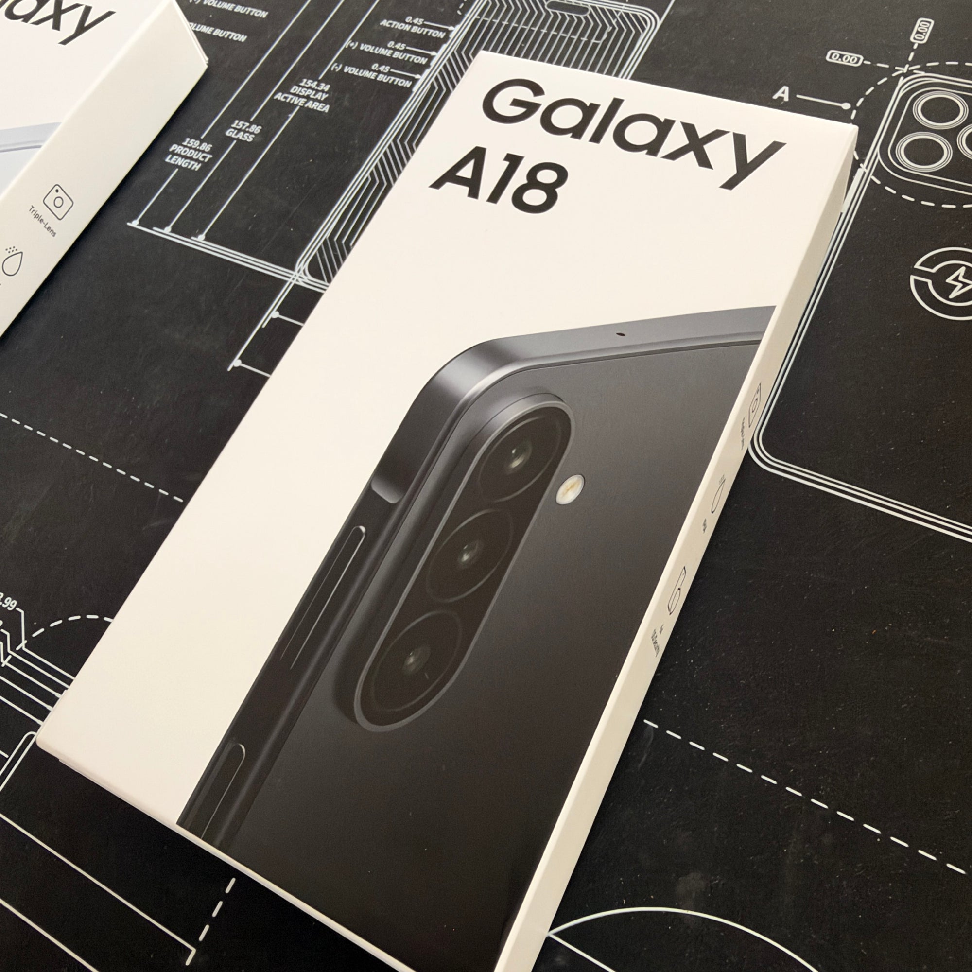 Samsung Galaxy A18 smartphone retail box shown at an angle on a black technical drawing surface. The front of the box is white with large black 'Galaxy A18' text and a dark phone image, and another box edge is partially visible at the top left.