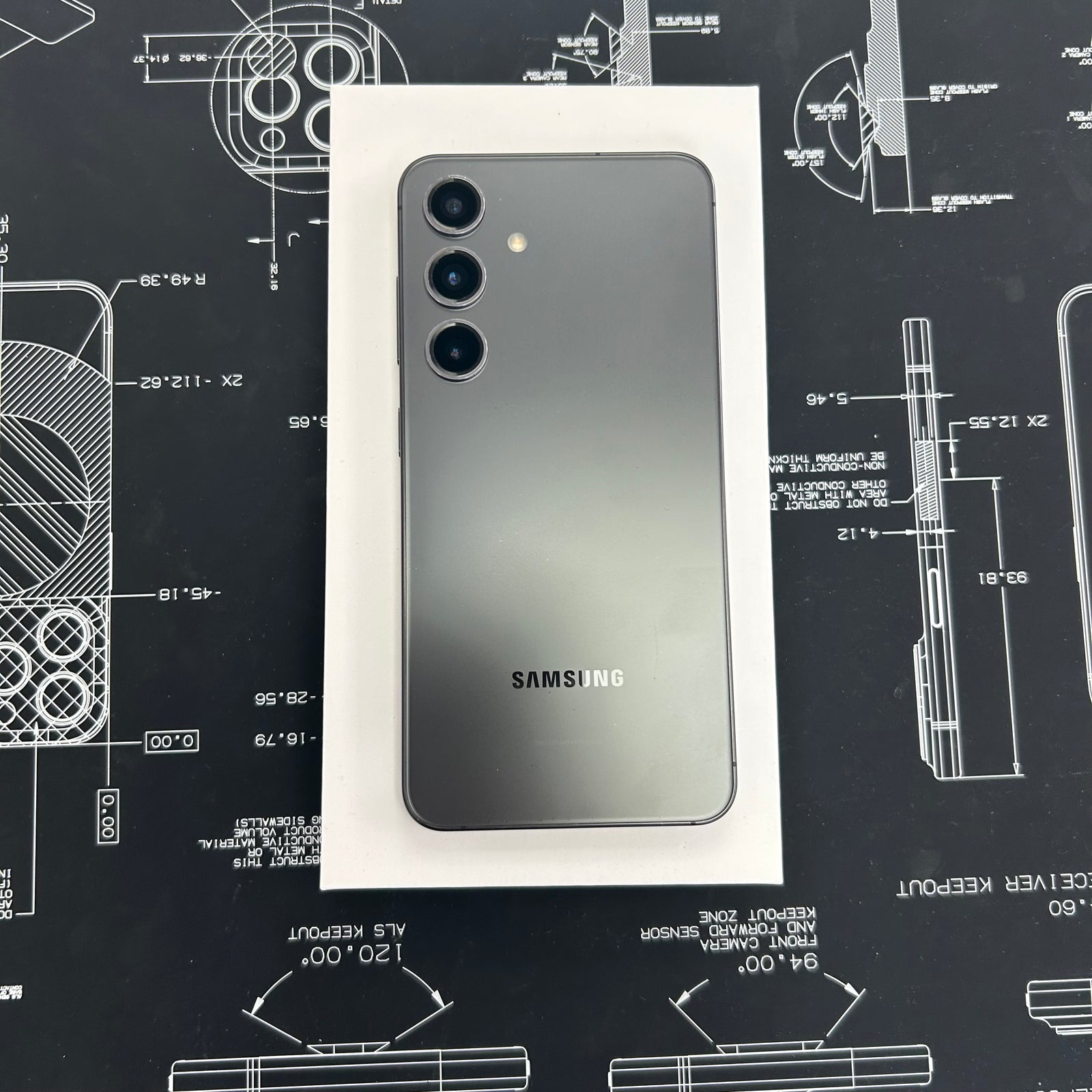 A Samsung S23+ smartphone shown from the back, resting on a white rectangular box over a dark technical blueprint surface. The phone has a dark gray to black gradient finish with three rear camera lenses and a visible SAMSUNG logo near the bottom.