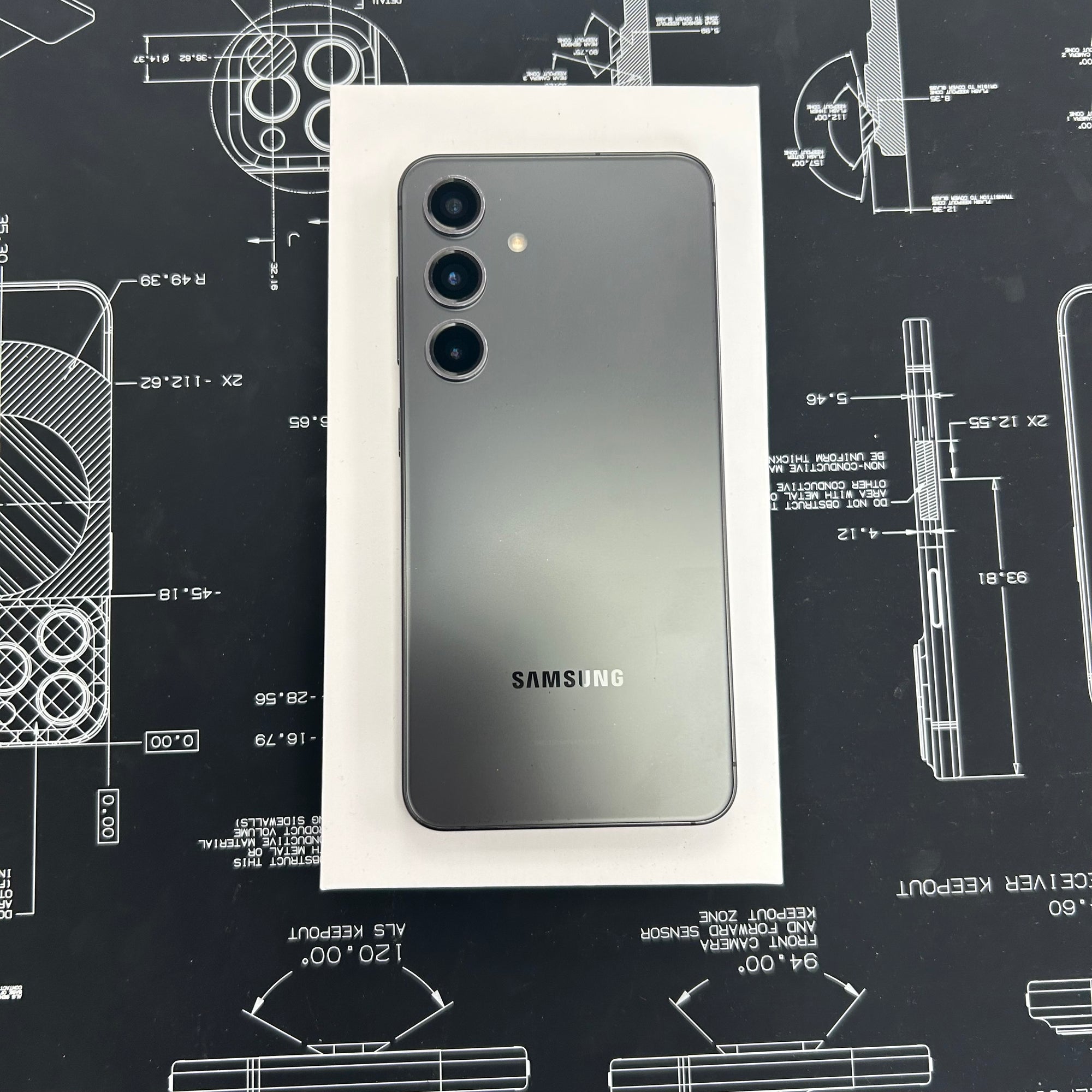 A Samsung S23+ smartphone shown from the back, resting on a white rectangular box over a dark technical blueprint surface. The phone has a dark gray to black gradient finish with three rear camera lenses and a visible SAMSUNG logo near the bottom.