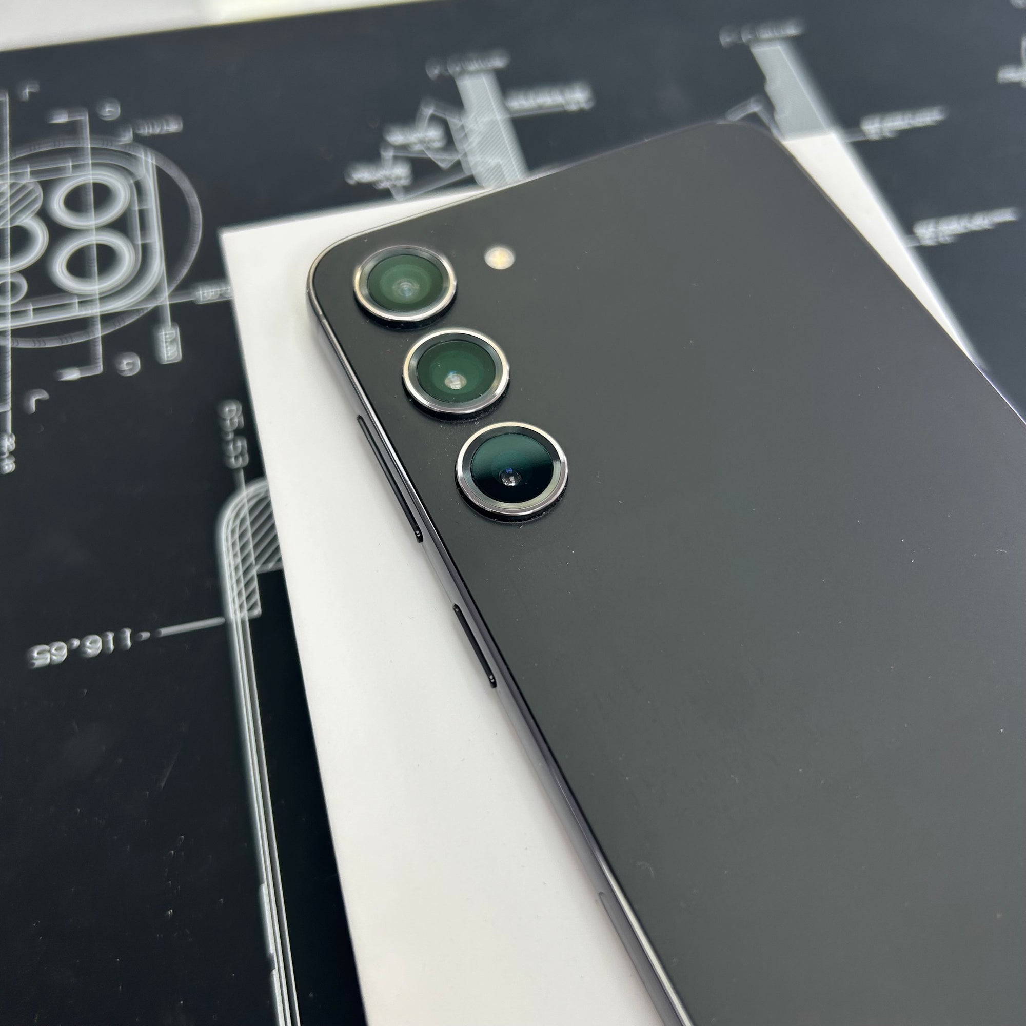 A close-up of a black S23+ smartphone showing the back of the device with three rear camera lenses, resting diagonally on a white sheet over a black technical blueprint background.
