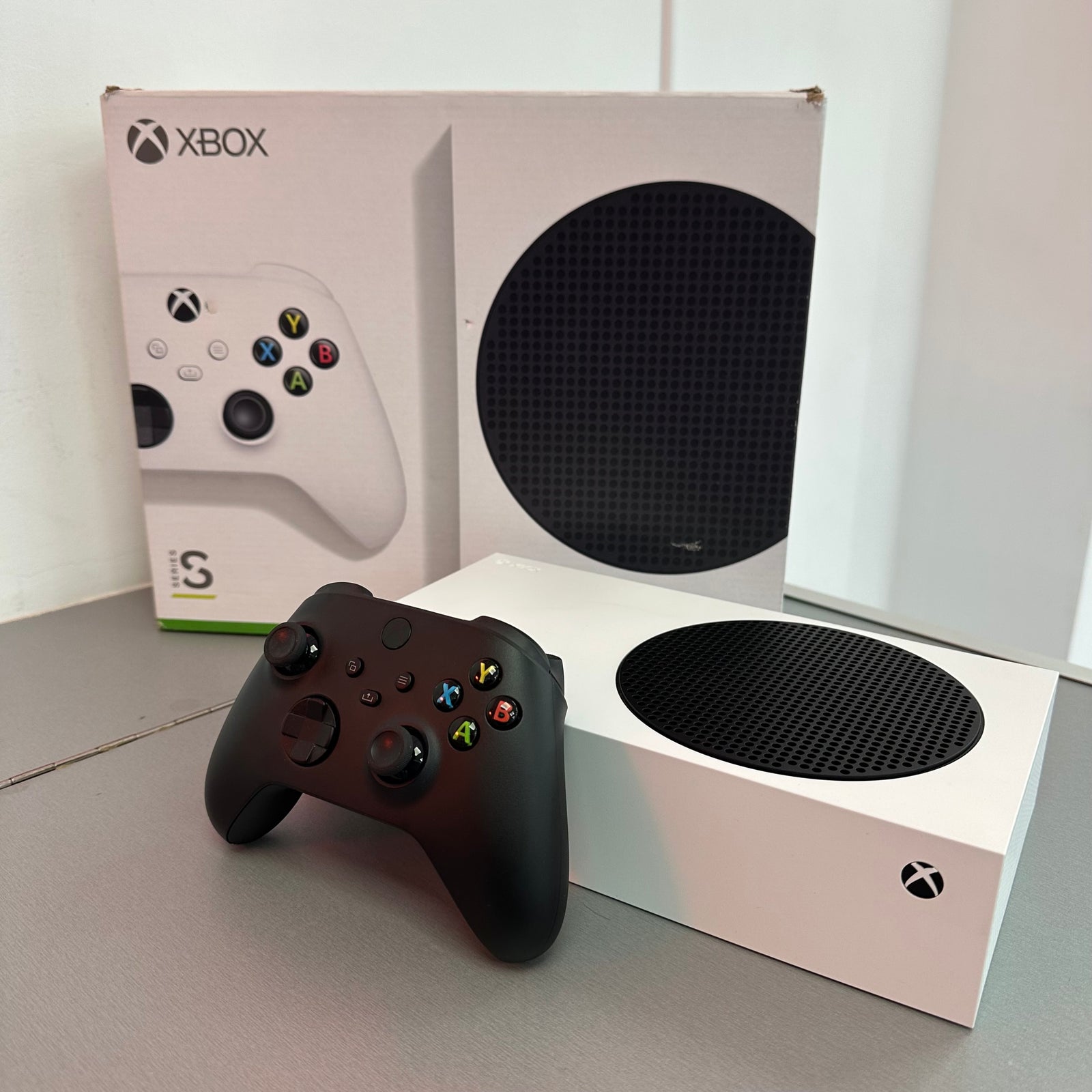 A white Xbox Series S console with a black textured skin, shown next to its black wireless controller and the original white product box on a gray tabletop.