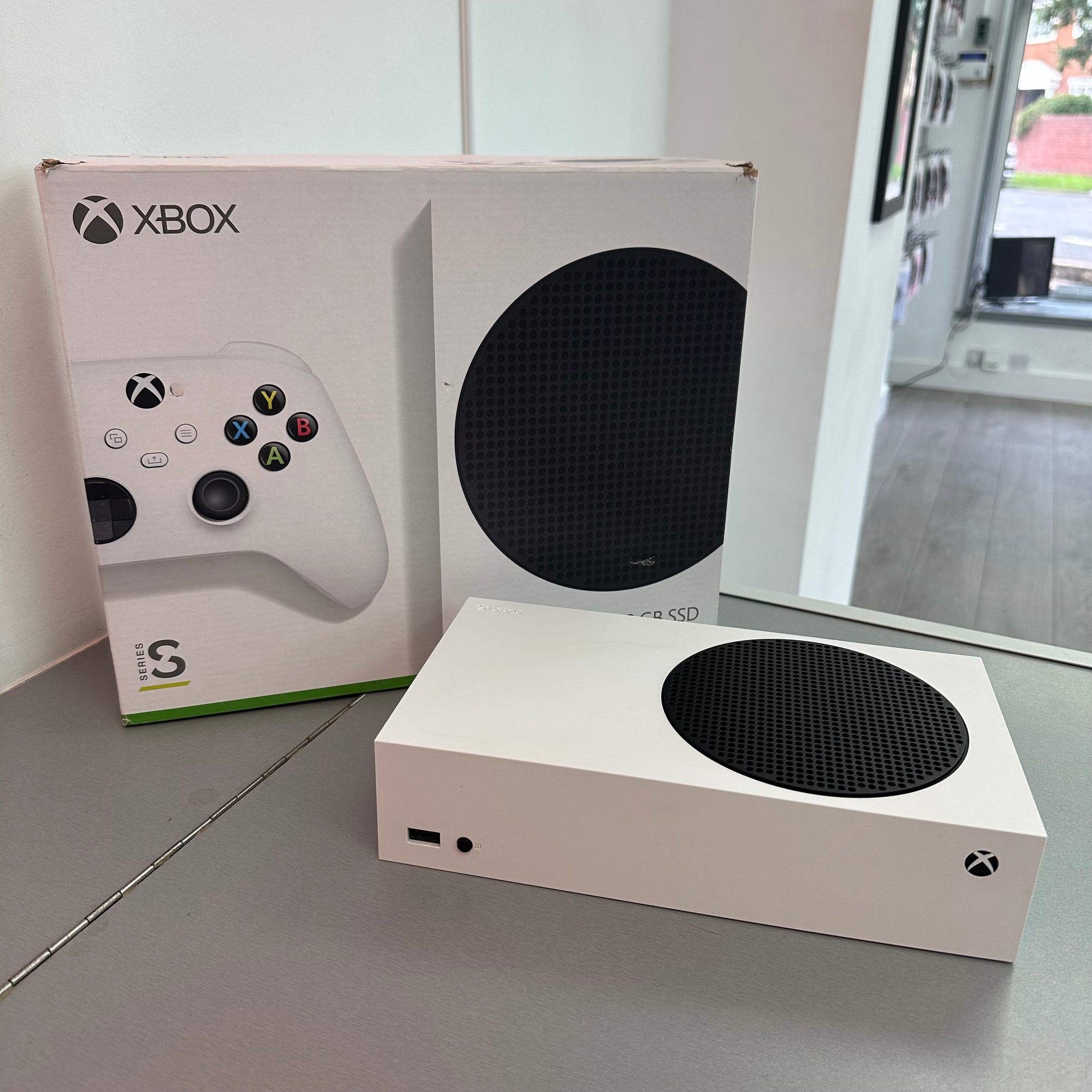 A white Xbox Series S console with a black circular vent on top, shown next to its original white retail box on a gray tabletop.