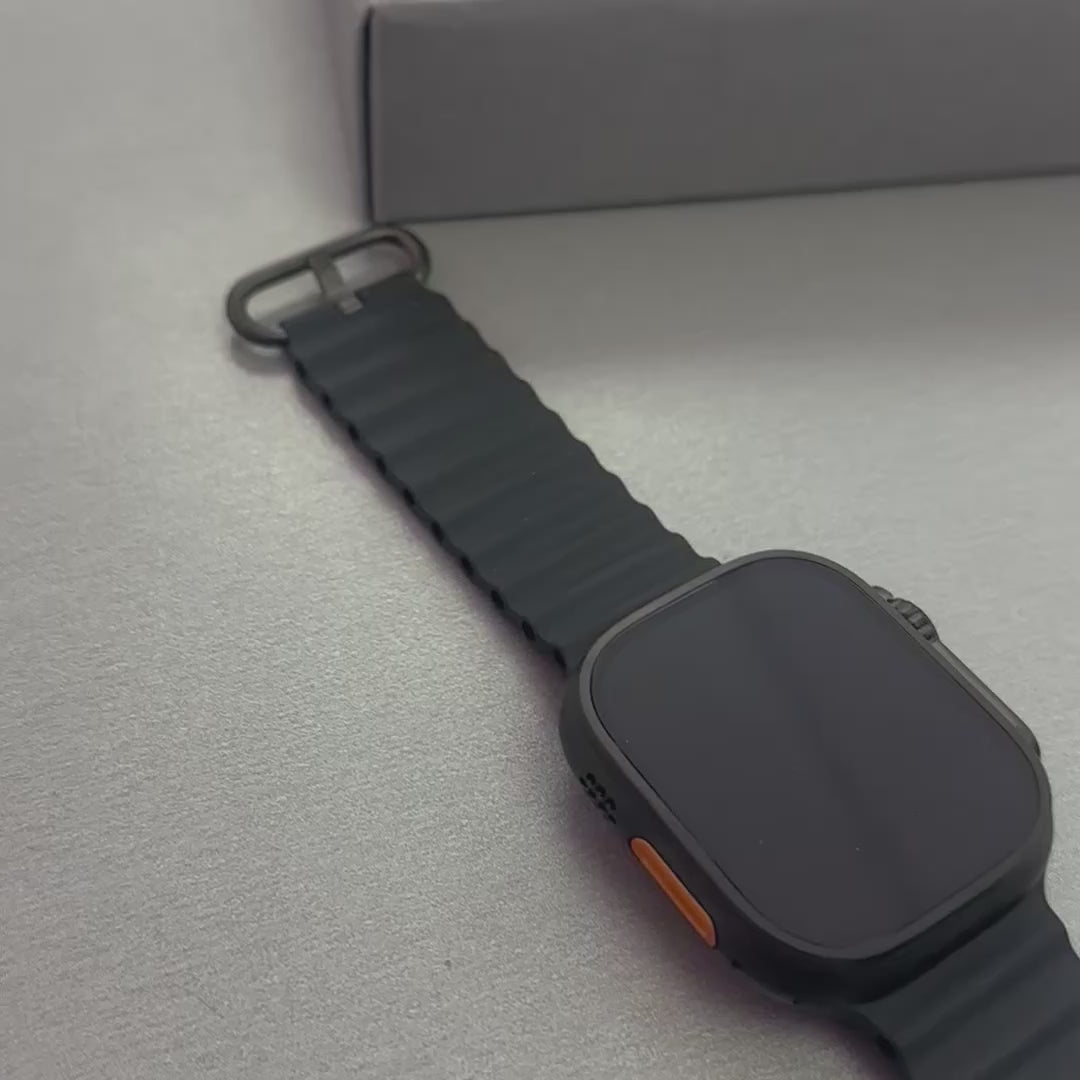 A black apple ultra smartwatch with a square screen and side button, resting in a gray foam insert tray inside a white box.