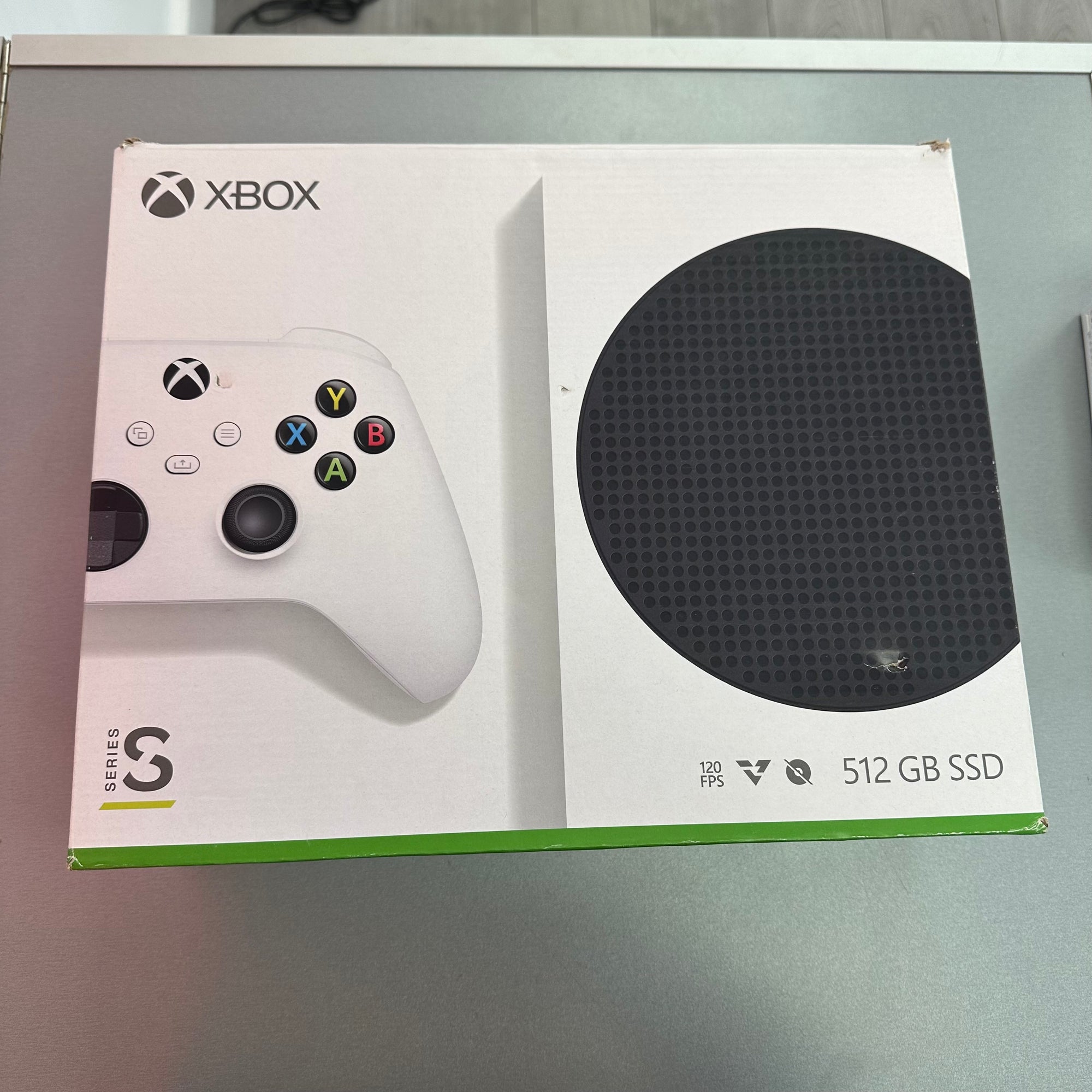 Xbox Series S 512 GB SSD console retail box in white, showing the controller and a black circular drive graphic on a gray tabletop.